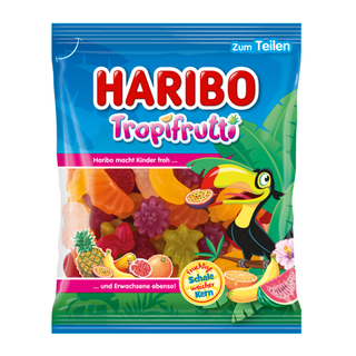 Produktbillede af en pose Haribo Tropifrutti frugtvingummier. Forsiden viser en farverig, tropisk designet emballage med blå baggrund, store grønne palmeblade og en tegnet tukan, der holder et åbent passionsfrugtstykke i næbbet. Teksten ‘HARIBO Tropifrutti’ står øverst i store røde og hvide bogstaver. I den gennemsigtige del af posen ses de mange frugtformede vingummier i forskellige farver som gul, rød, orange, grøn og lilla. Nederst er illustreret tropiske frugter såsom ananas, banan, figen og pink grapef
